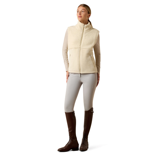 A woman modeling a cream sherpa fleece vest over a beige long-sleeve shirt, paired with light gray riding breeches and dark brown riding boots, against a plain white background.