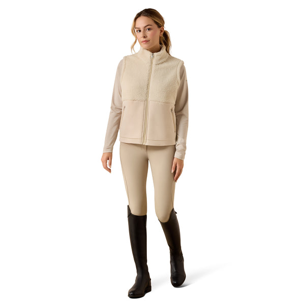 A woman modeling a light beige fleece zip-front vest and matching beige riding tights with black riding boots, photographed against a clean white studio background.