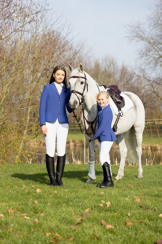 Two people in equestrian attire standing next to a white horse in a grassy field.