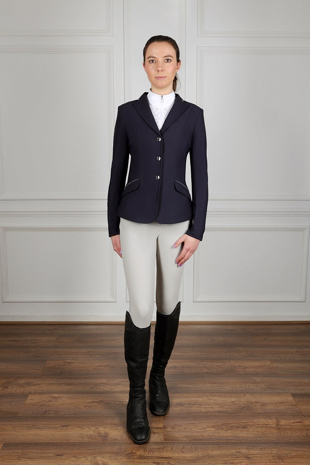 Person wearing a navy riding jacket, white shirt, beige riding pants, and black riding boots in a room with wooden floor and white paneled walls.