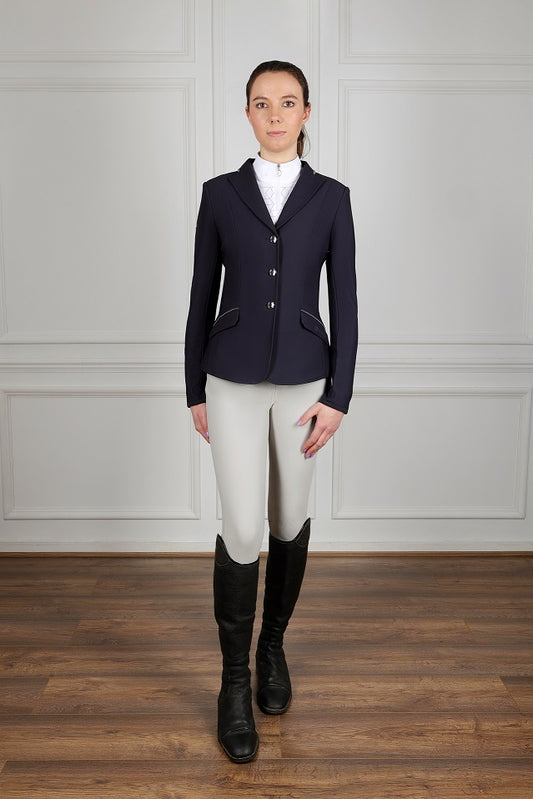Person wearing a navy riding jacket, white shirt, beige riding pants, and black riding boots in a room with wooden floor and white paneled walls.