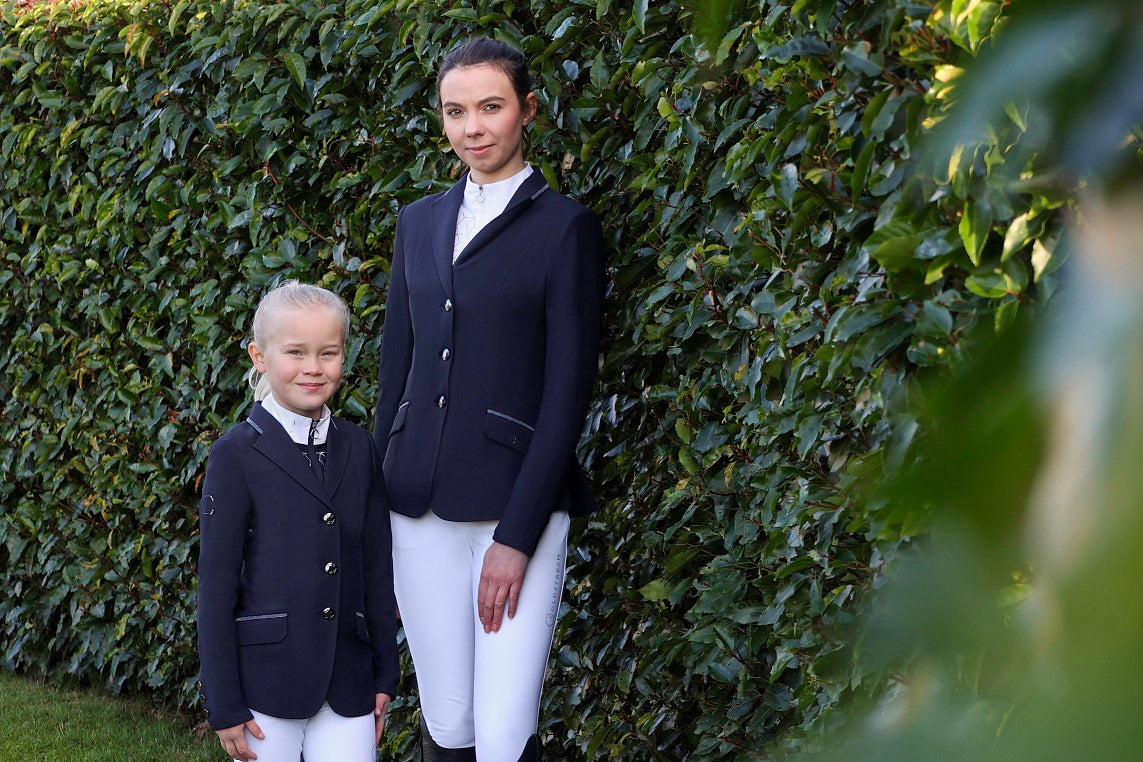Two girls in navy competition jackets and riding gear in an outdoor setting.