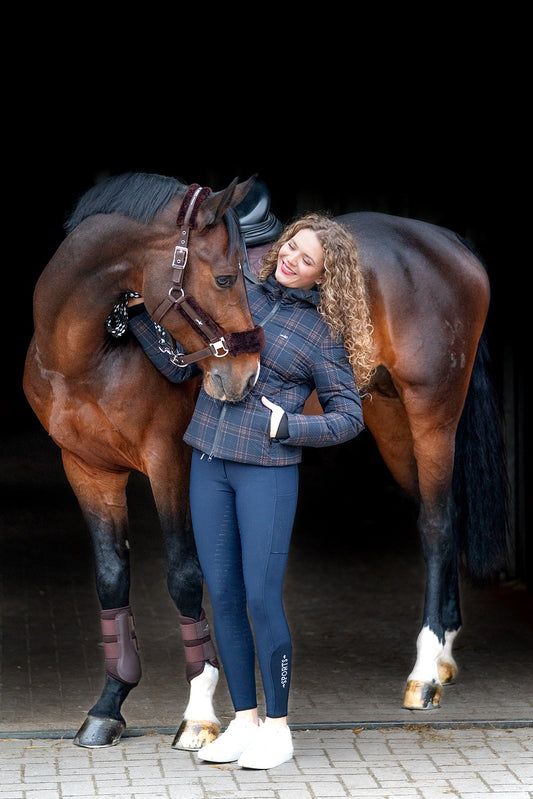 A woman in equestrian apparel stands smiling next to a brown horse in a stable doorway. The woman wears a blue plaid jacket, dark blue riding tights, white riding boots, and a black riding helmet.
