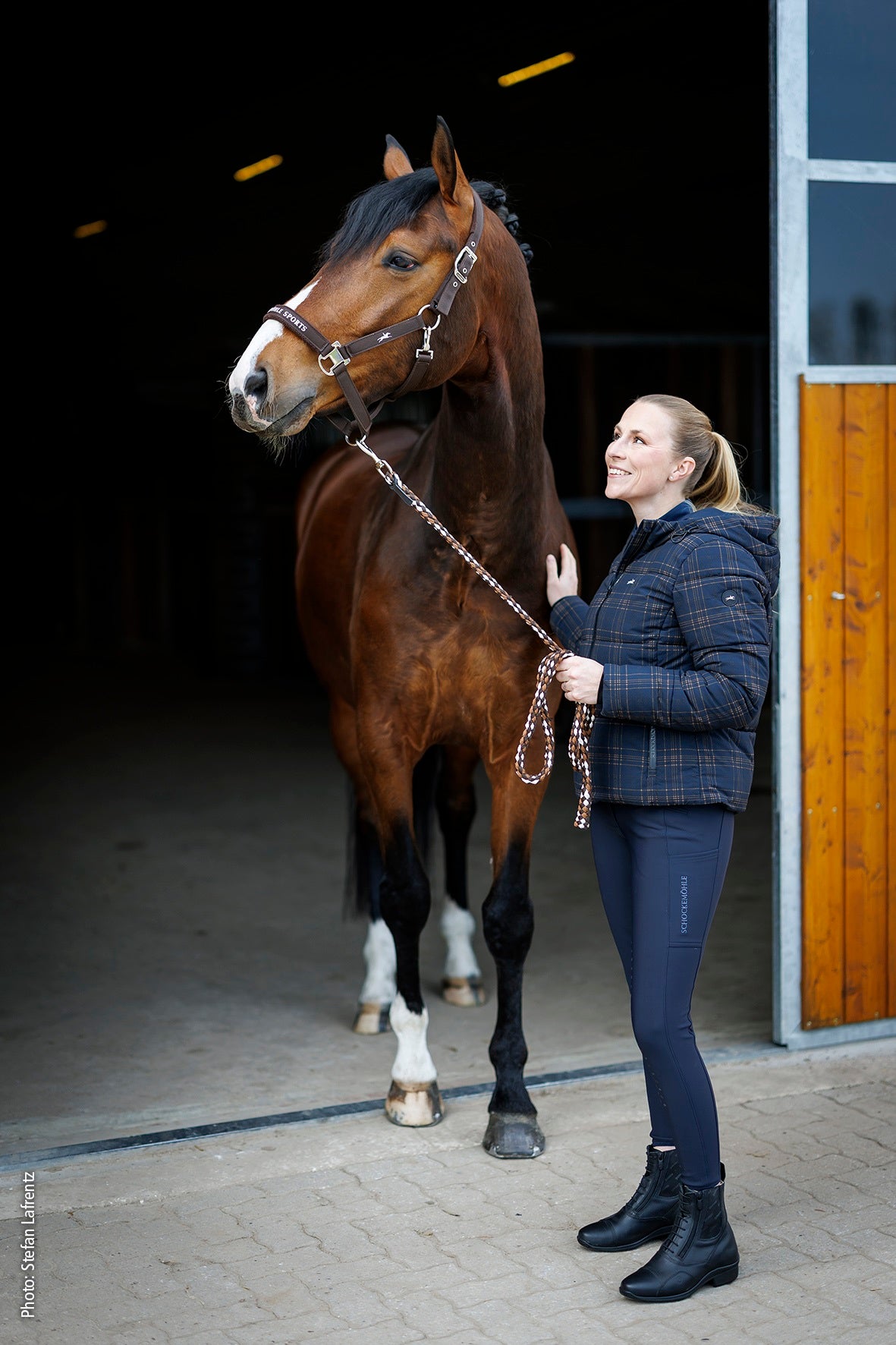 A woman in a dark blue plaid jacket and leggings stands beside a brown horse with white socks, holding a lead rope in front of a stable doorway.