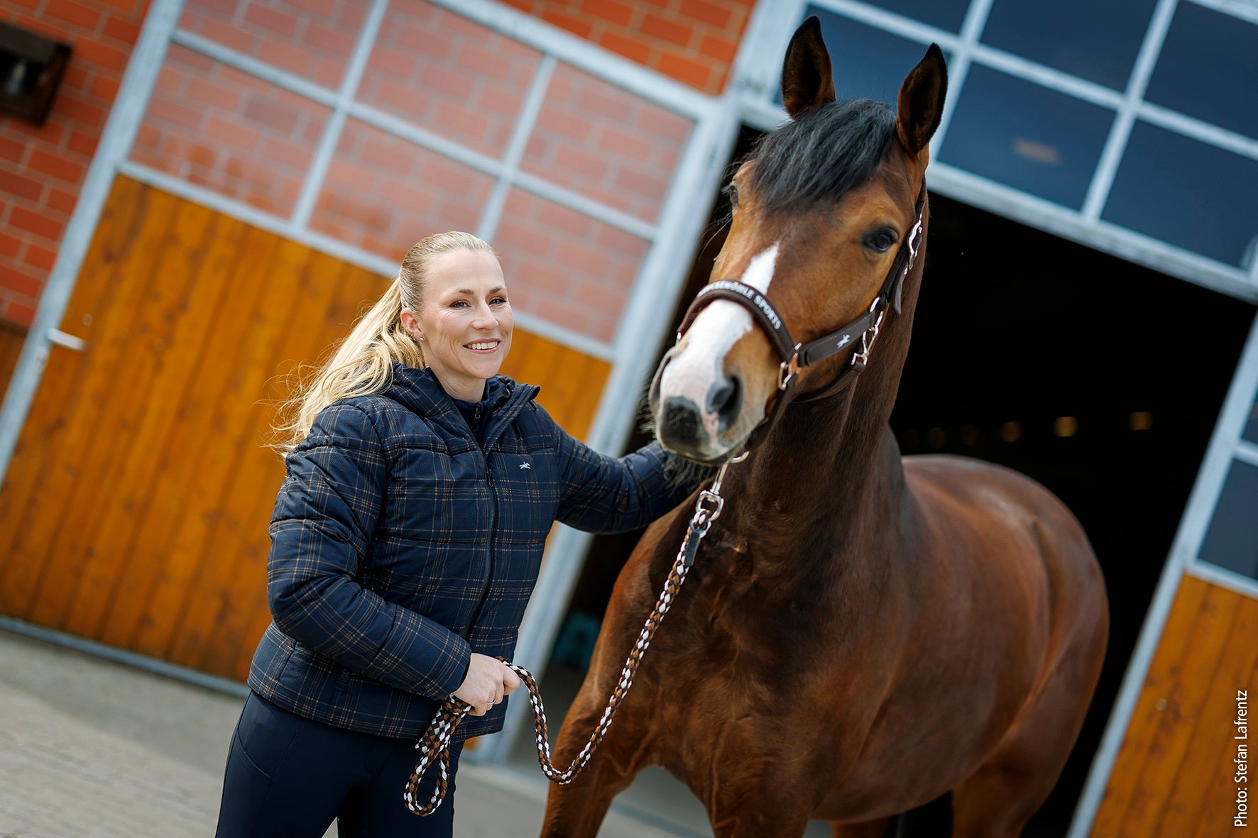 A woman in a dark blue plaid puffer jacket stands beside a brown horse with a white blaze, holding its lead rope in front of a stable with orange-brown wooden doors and a red brick wall.