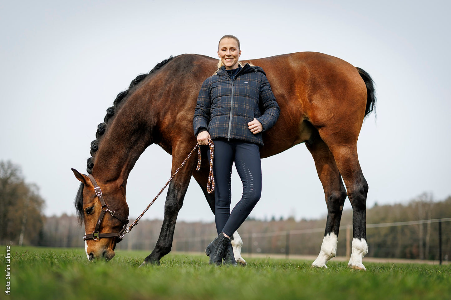 A woman stands outdoors beside a large brown horse grazing in a grassy field, holding a lead rope. She is wearing a dark quilted puffer jacket, dark leggings, and black boots.