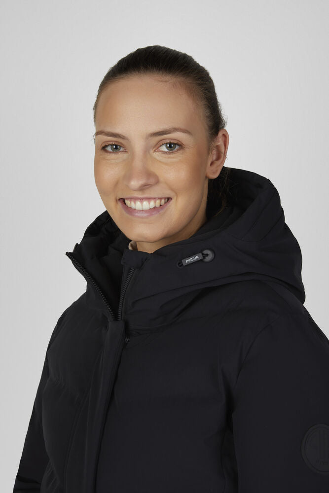 A smiling woman with her hair pulled back wears a black hooded winter jacket against a light gray studio background.