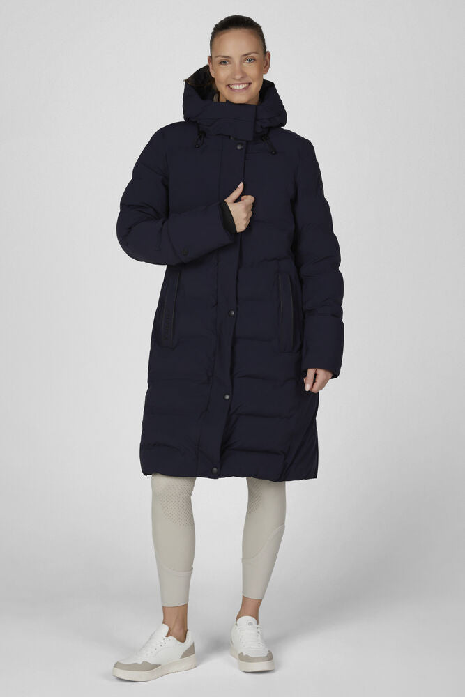 A woman modeling a long navy blue puffer coat with a high hood, paired with light beige leggings and white sneakers, against a light gray studio background.