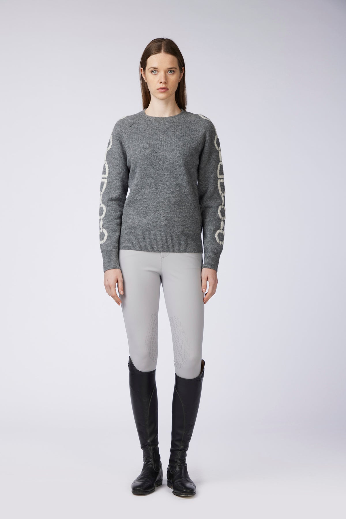 A model stands against a light gray studio background wearing a heather gray crewneck sweater with white circular chain-link graphics down both sleeves, light gray fitted pants, and black riding boots.