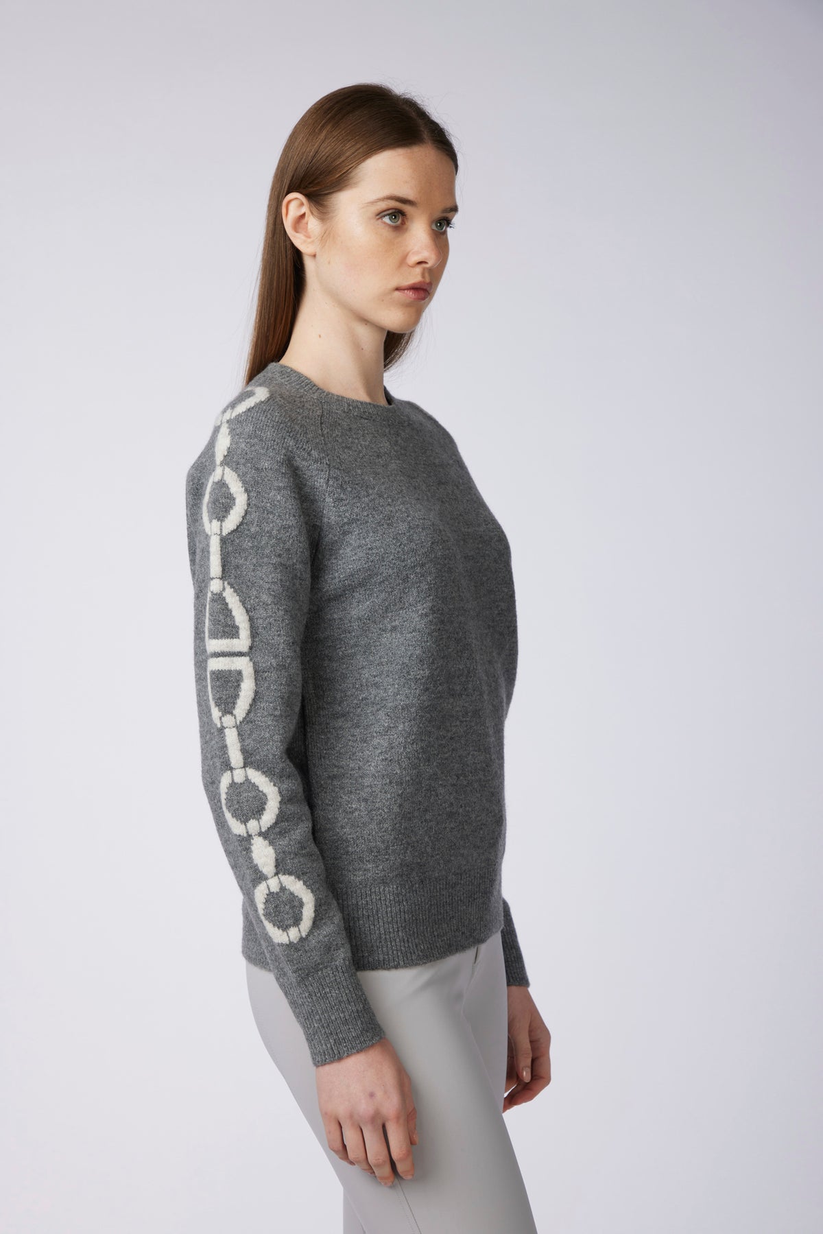 A woman wearing a gray knit sweater with a cream-colored chain motif down one sleeve, paired with light gray pants, photographed against a plain light gray studio background.