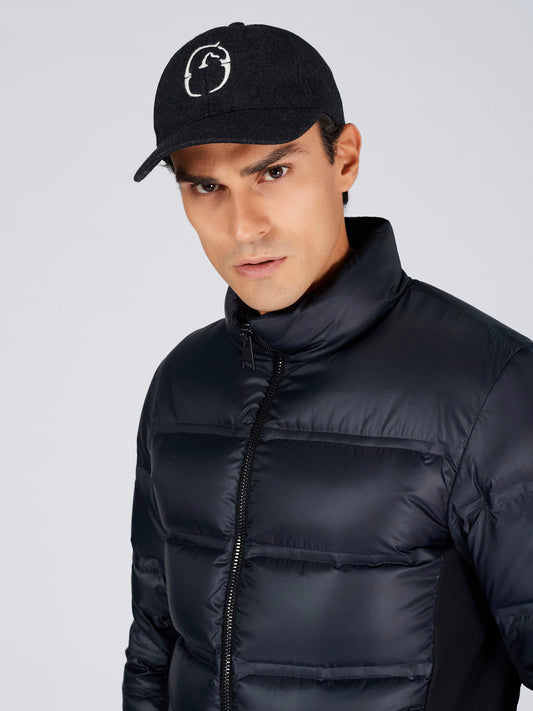 A man wearing a navy puffer jacket with a high collar and a black baseball cap featuring a white circular logo, photographed against a light gray studio background.