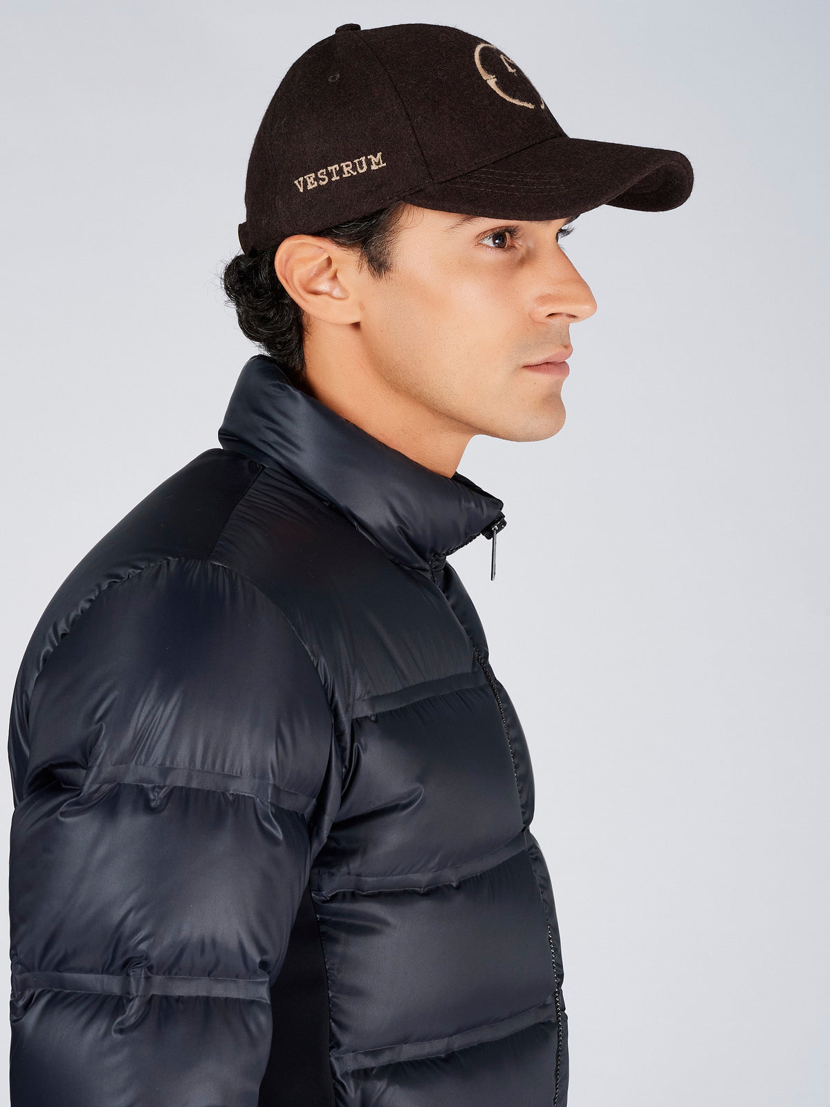 A man in profile wearing a black puffer jacket and a black baseball cap with 'VESTRUM' embroidered on the side, photographed against a light gray studio background.
