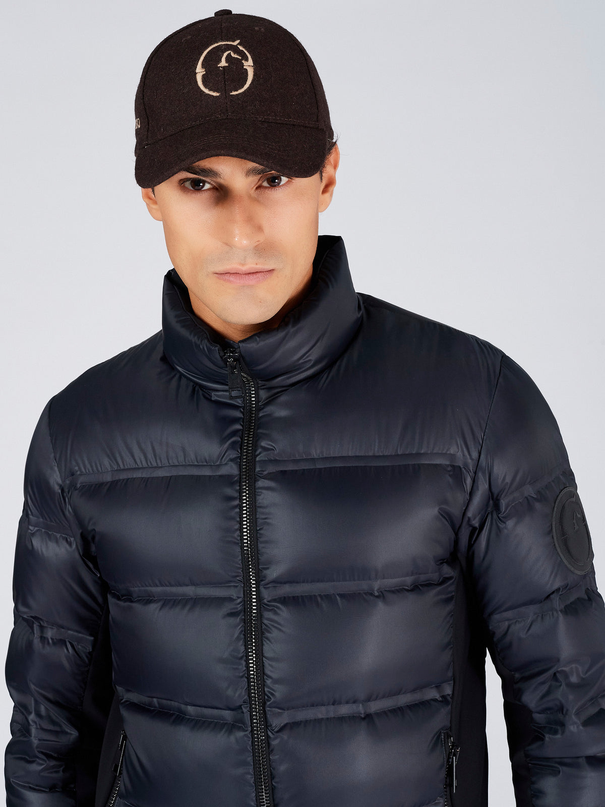 A man wearing a dark navy puffer jacket with a high collar and a dark brown knit cap featuring a circular logo, photographed against a light gray studio background.