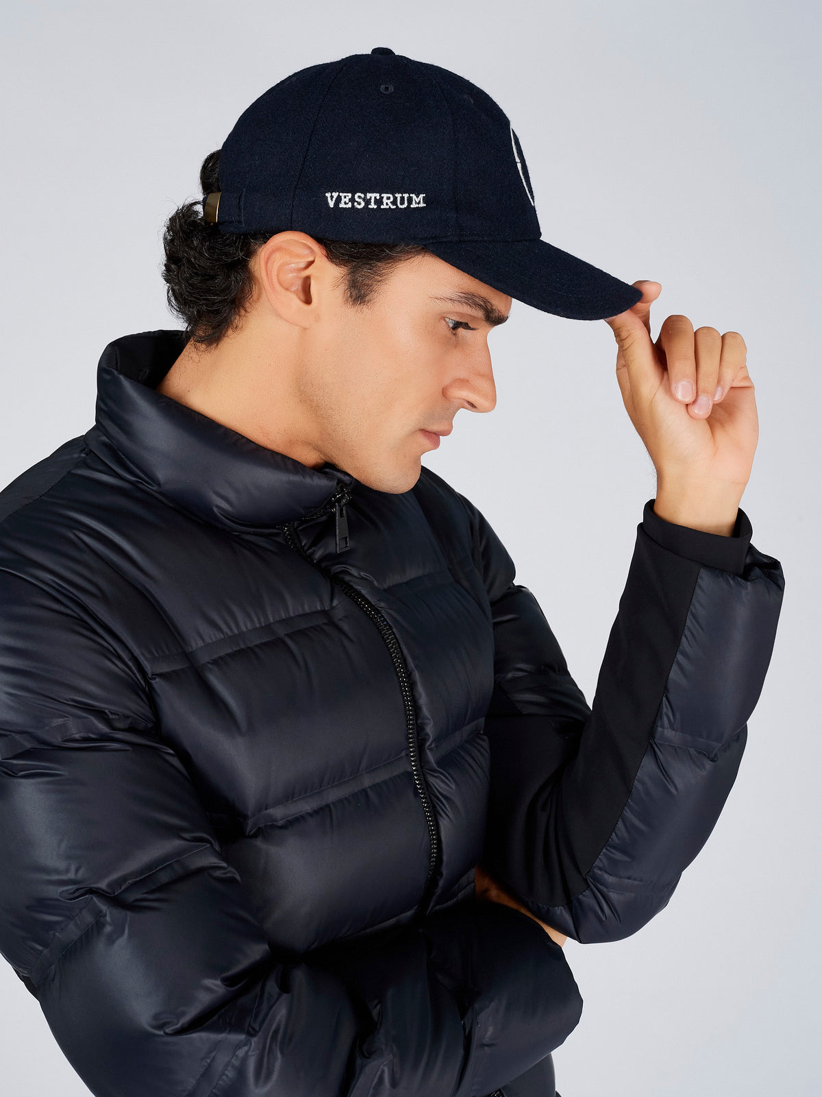 A man wearing a black puffer jacket and a navy Vestrum baseball cap, posing against a light gray studio background.