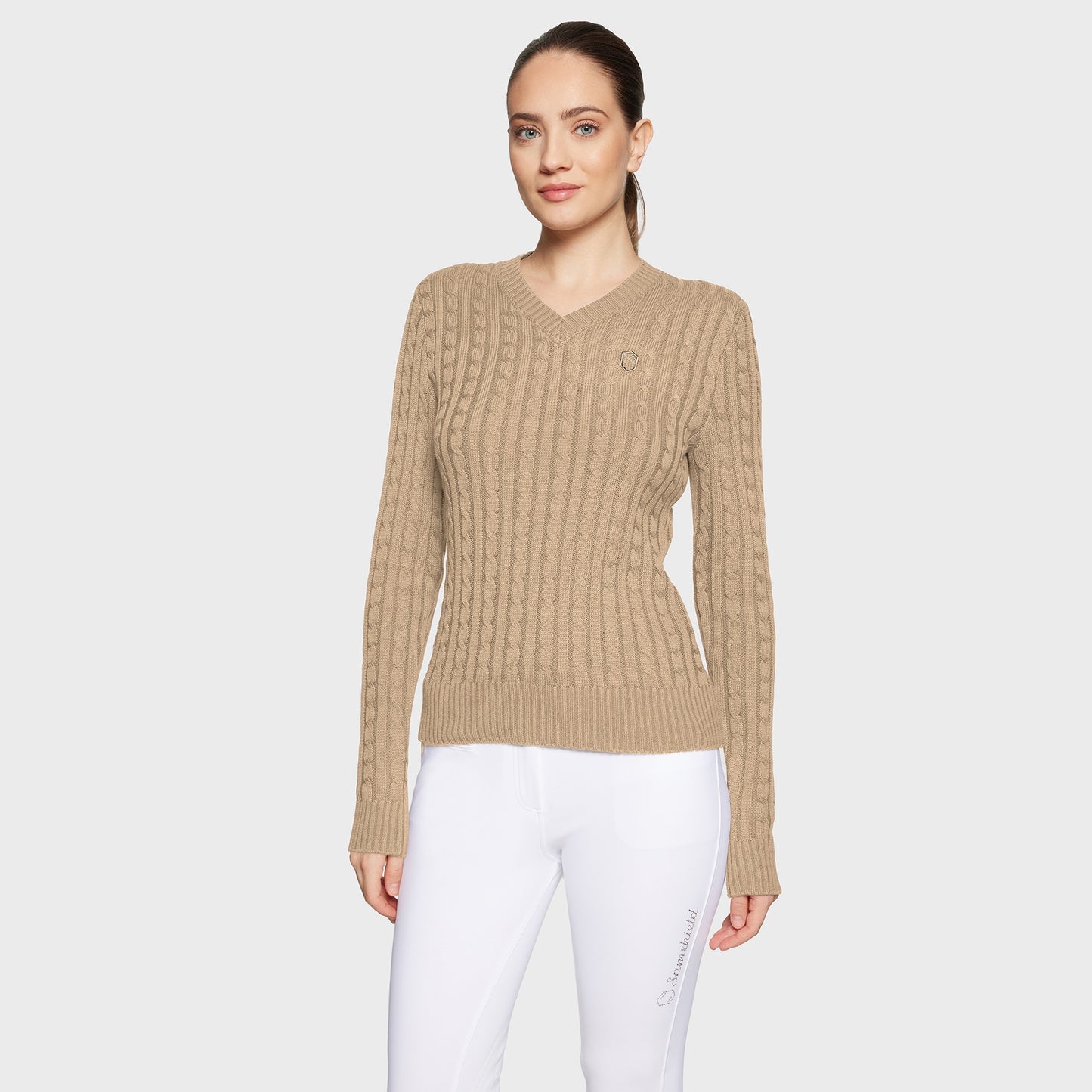 Woman wearing a beige cable knit sweater and white pants on a light gray background