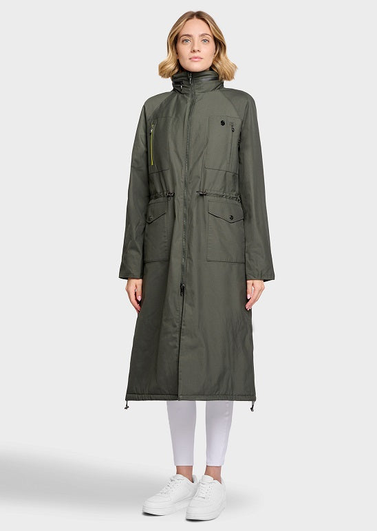 A woman modeling a long olive green hooded parka coat with front flap pockets and drawstring details, paired with white leggings and white sneakers, against a light gray studio background.
