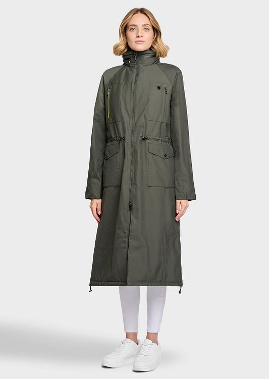 A woman modeling a long olive green hooded parka coat with front flap pockets and drawstring details, paired with white leggings and white sneakers, against a light gray studio background.