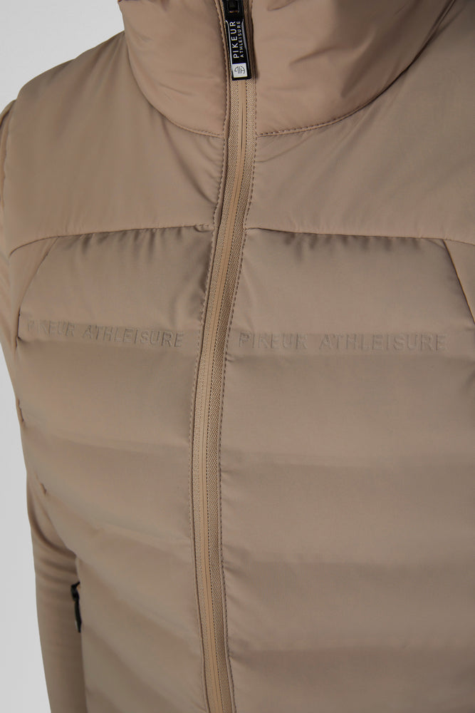 Close-up of a beige puffer vest with a high collar and front zipper, featuring subtle tonal branding text across the chest.