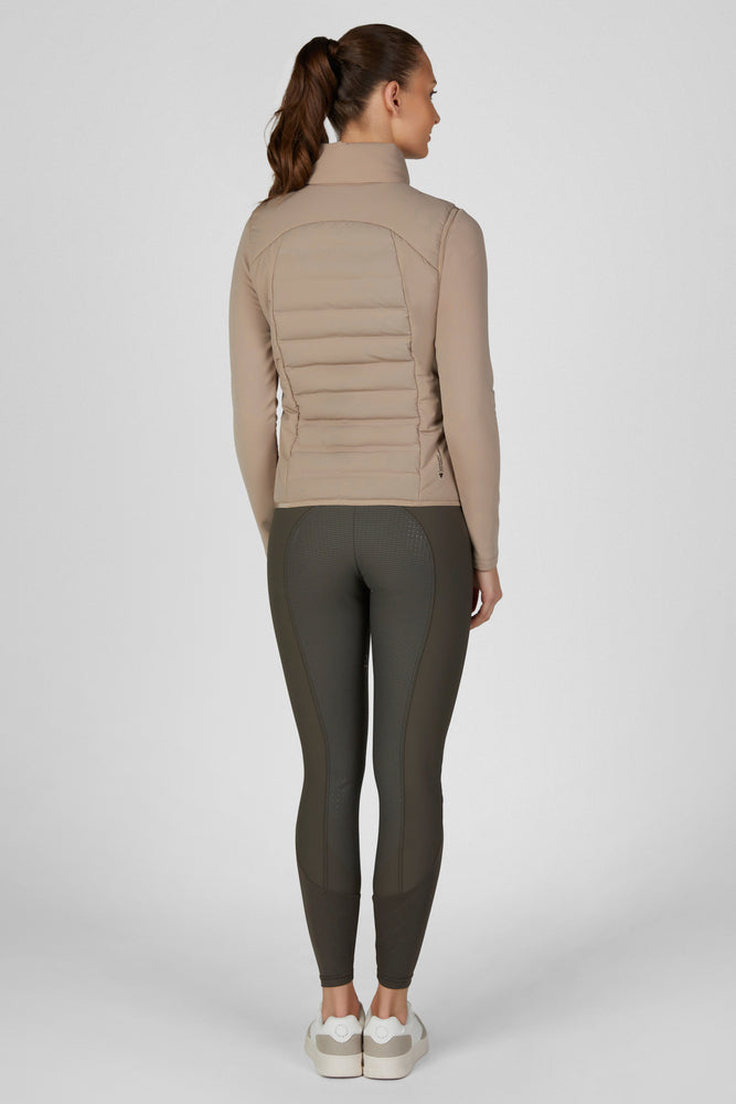 A woman modeling a light brown puffer vest over a matching long-sleeve top and dark olive leggings, paired with white sneakers, against a light gray studio background.