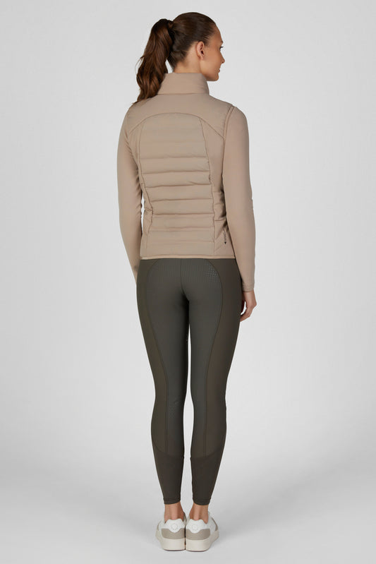 A woman modeling a light brown puffer vest over a matching long-sleeve top and dark olive leggings, paired with white sneakers, against a light gray studio background.