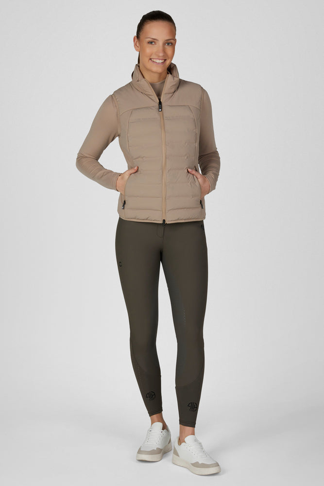 A woman modeling a light brown puffer vest over a matching long-sleeve top and dark olive leggings, paired with white sneakers, against a light gray studio background.