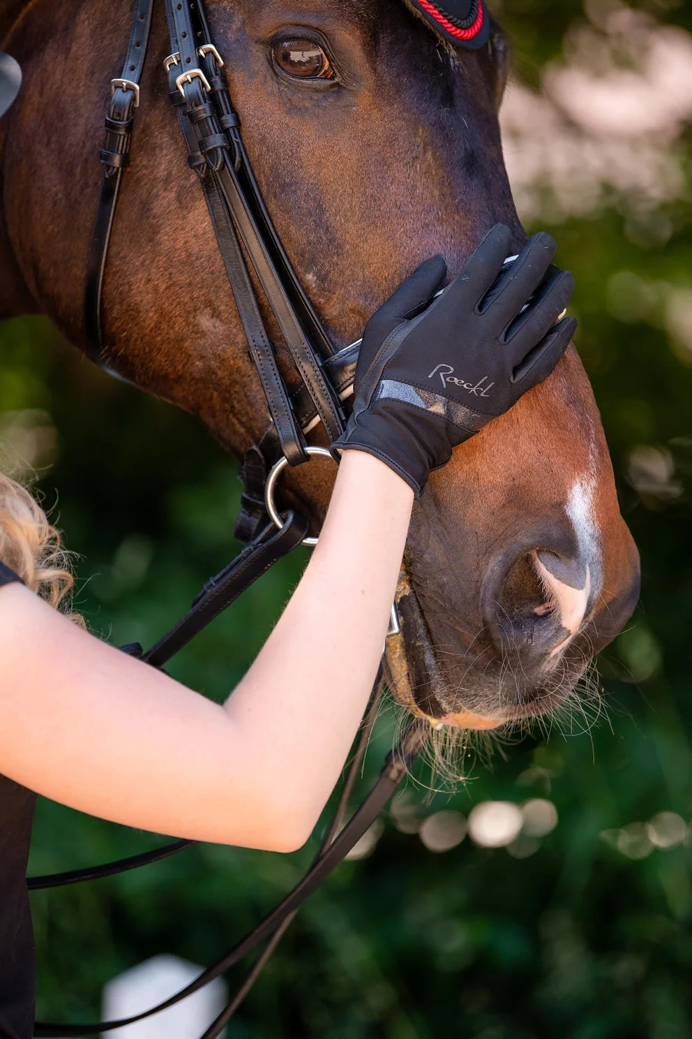 Black equestrian riding gloves with a white 'Rouge' logo, worn by a person touching a brown horse in an outdoor setting.