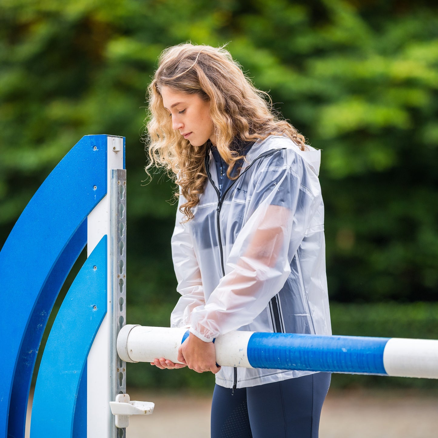 Girl putting up a blue showjumping pole wearing a transparent rain jacket 