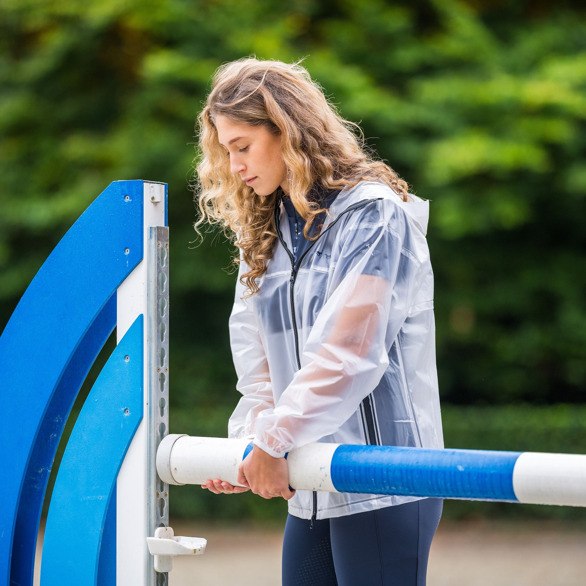 Girl putting up a blue showjumping pole wearing a transparent rain jacket 