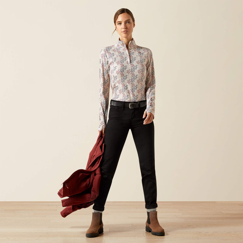 Woman wearing a patterned shirt, black pants, and brown boots holding a red jacket against a plain background