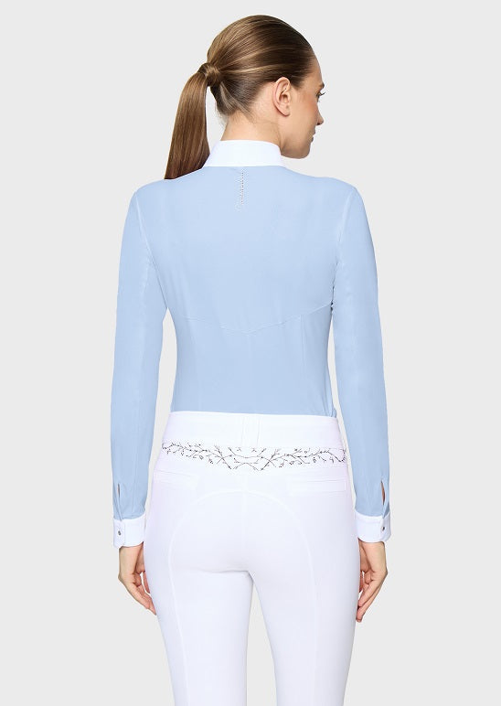 A woman shown from the back wearing a light blue long-sleeve equestrian-style top with a white collar and white fitted pants with decorative embroidery at the waist, against a light gray studio background.