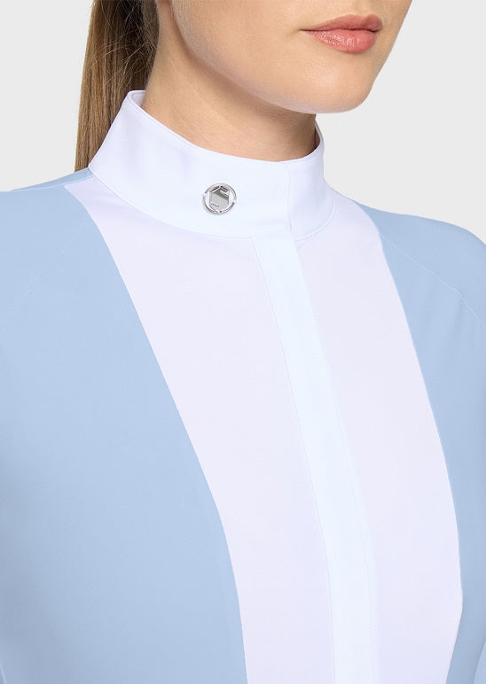 A woman wearing a light blue long-sleeve equestrian-style top with a white collar against a light gray studio background.