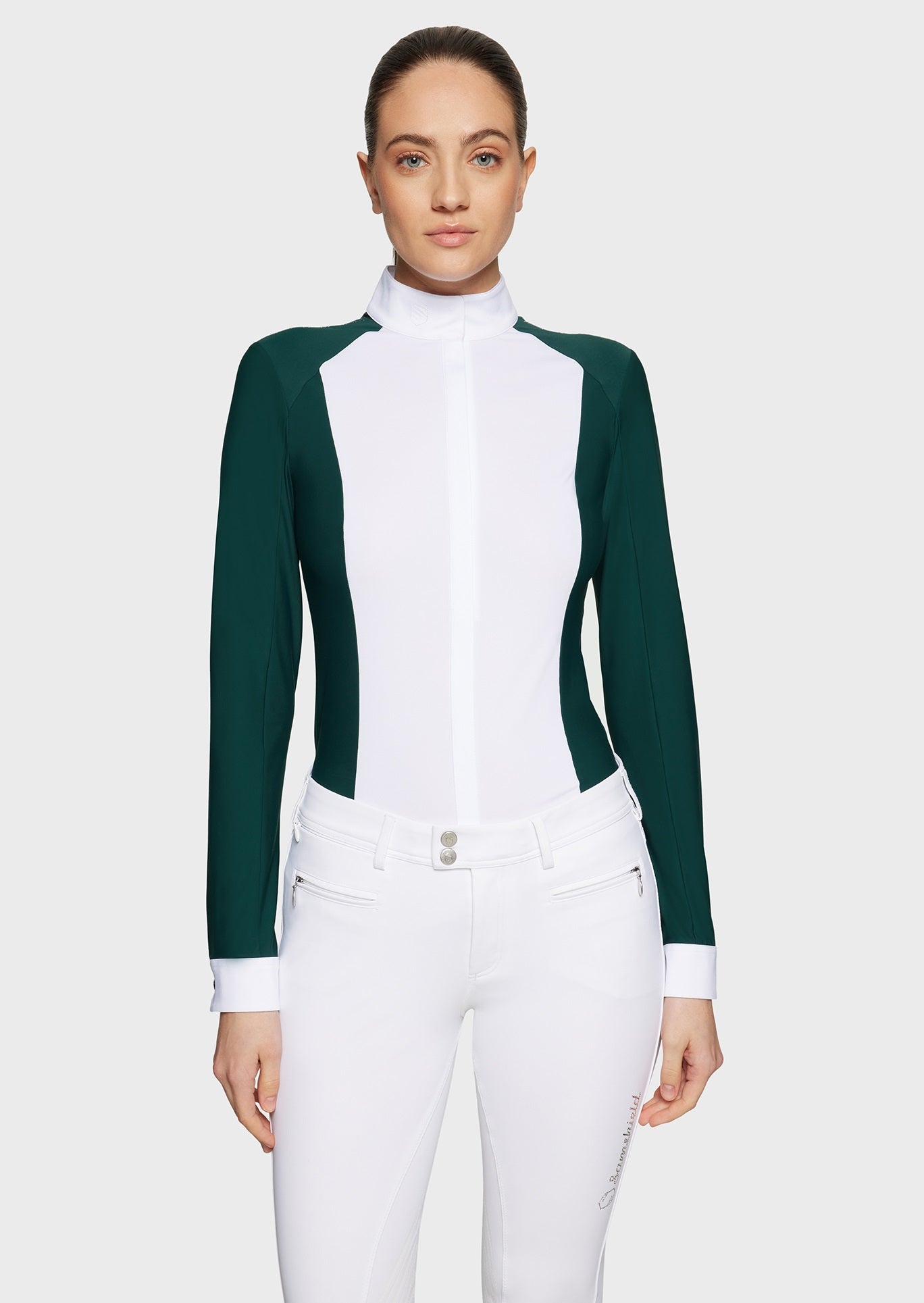 Woman wearing a green competition shirt with a white bib and white breeches on a beige background