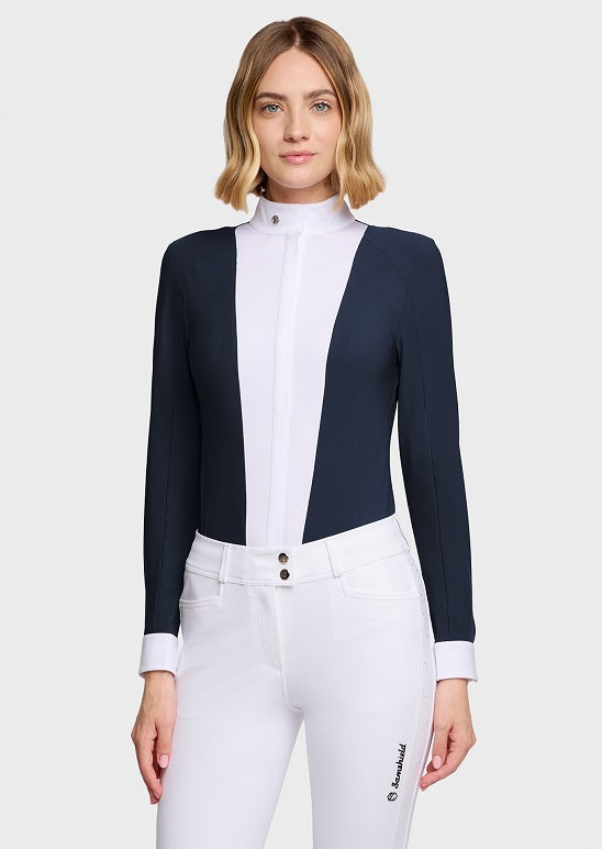 A woman modeling a long-sleeve equestrian-style jacket with a high mock neck, featuring navy blue side panels and a white center panel, paired with white riding pants against a light gray studio background.