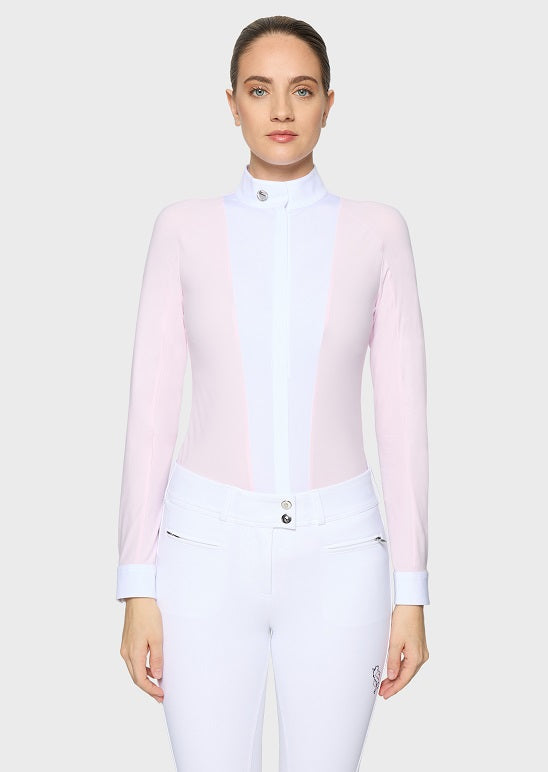 A woman modeling a light pink and white long-sleeve equestrian-style mock neck top paired with white riding breeches, against a solid light gray background.