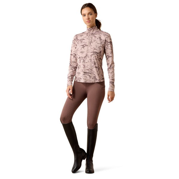 A woman modeling a long-sleeve equestrian-style top with a dusty pink and taupe floral pattern, paired with brown leggings and black riding boots, against a plain white background.
