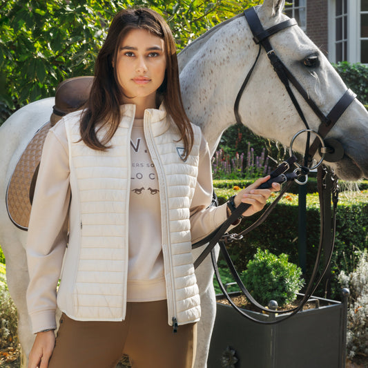 Girl wearing a cream gilet and brown breeches holding a grey horse with a house in the background