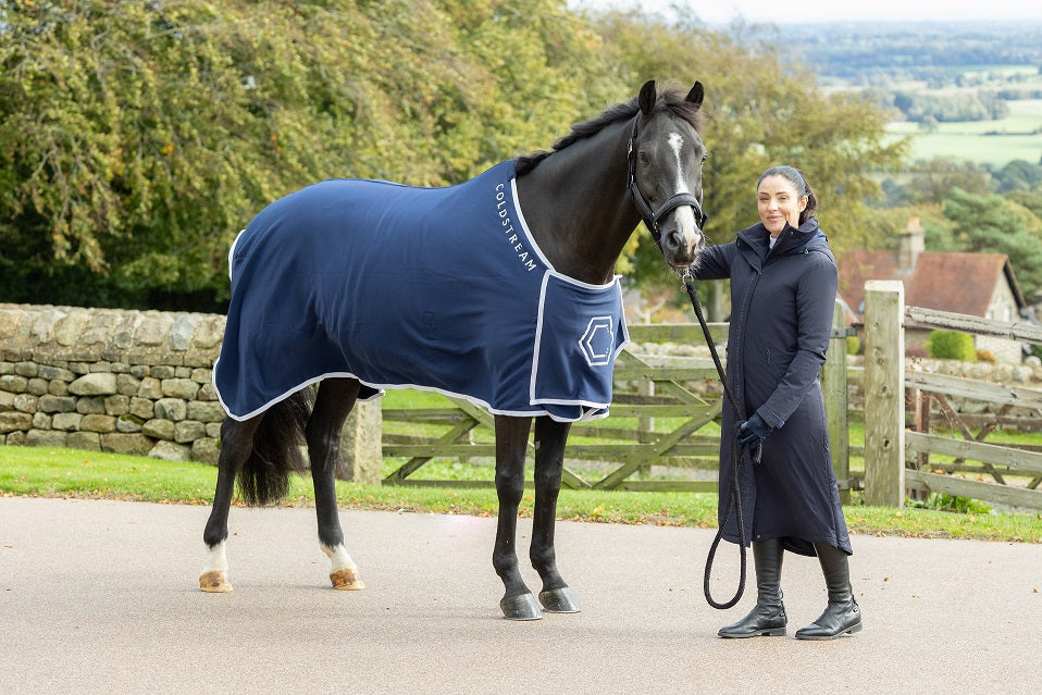 Woman in a dark coat standing next to a horse outdoors with trees and a fence in the background.