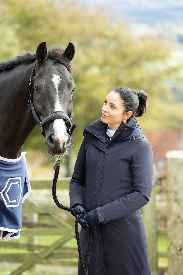 Woman in a dark coat standing next to a horse outdoors with trees and a fence in the background.