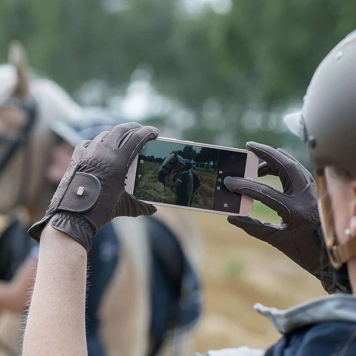 A close-up lifestyle shot of a person wearing brown leather equestrian gloves and a light gray riding helmet, holding a smartphone to take a photo of a horse in a field. Another horse rider is blurred in the background.