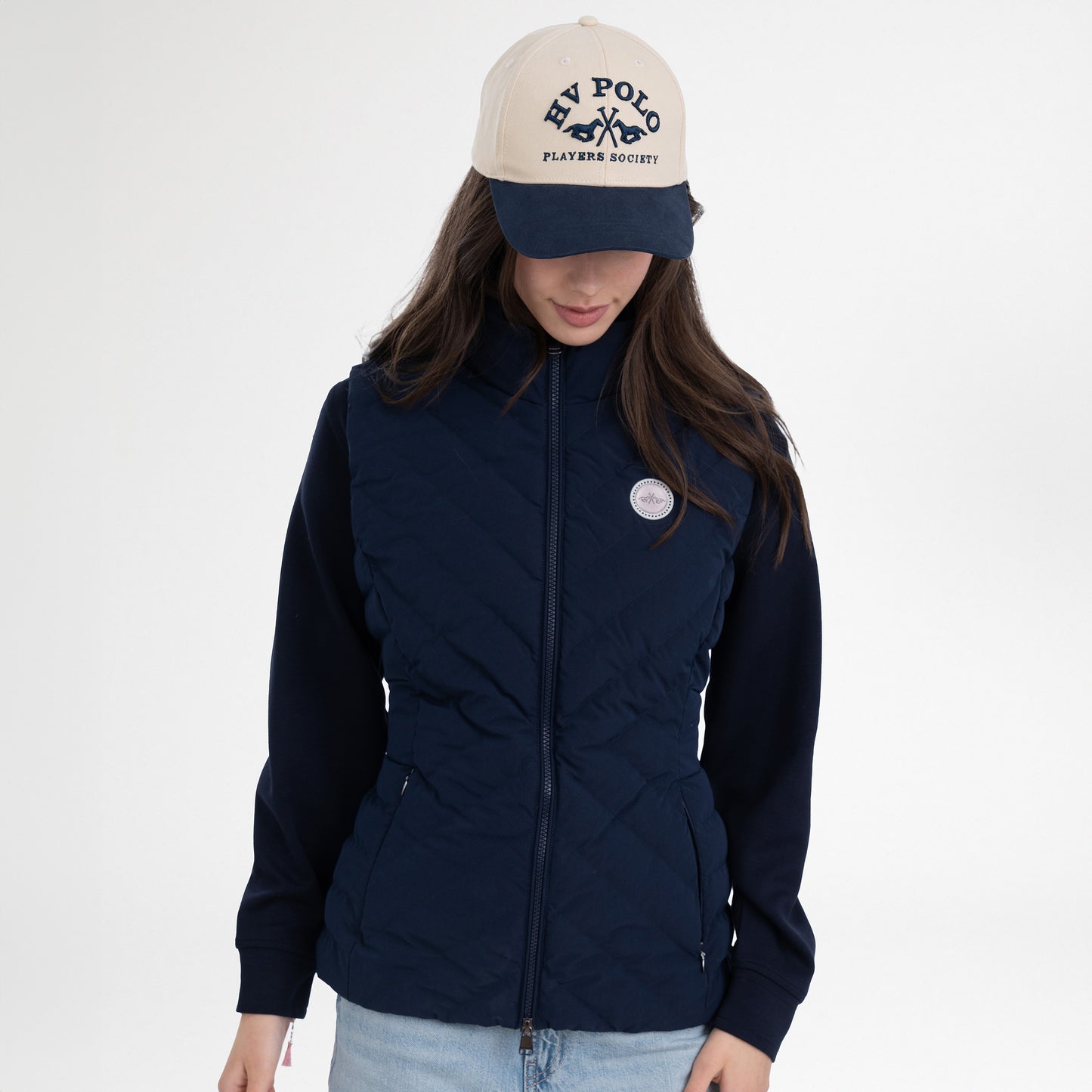 Girl wearing a navy gilet, navy hoodie, navy and beige cap and blue jeans on a white background