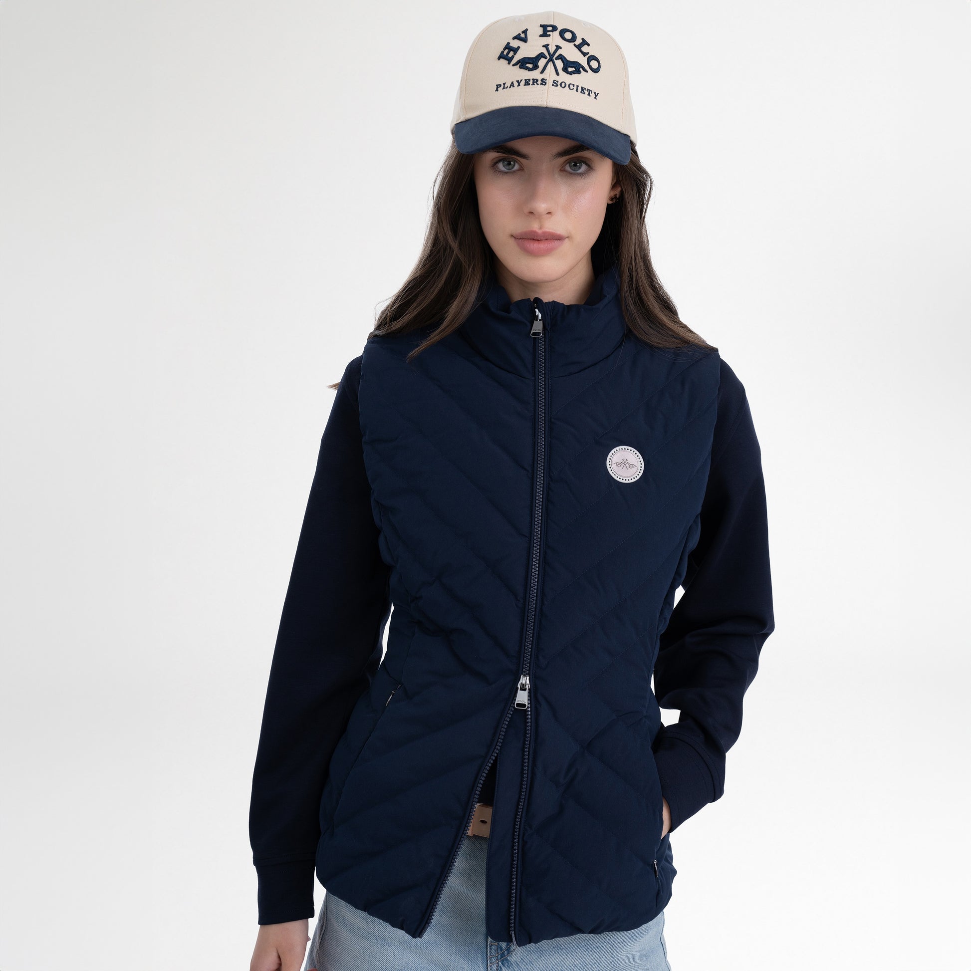 Girl wearing a navy gilet, navy hoodie, navy and beige cap and blue jeans on a white background