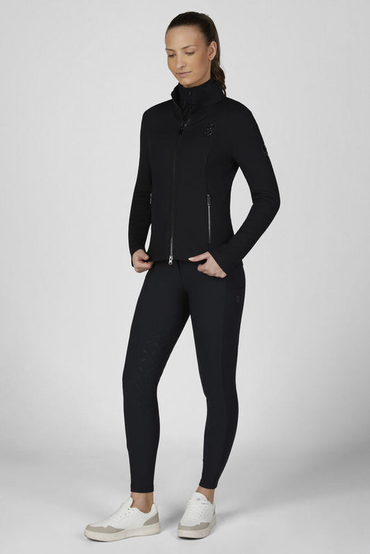 A woman modeling a black long-sleeve athletic zip-up jacket and matching black leggings with white sneakers, standing against a light gray studio background.