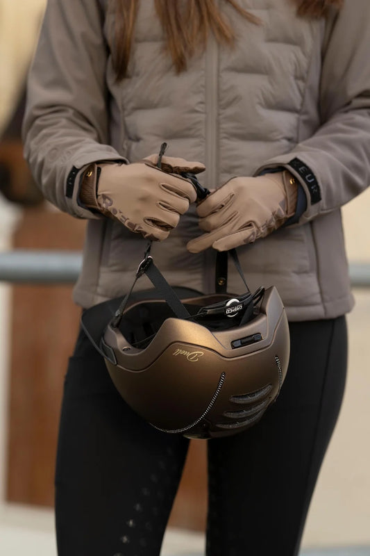 A person wearing a taupe quilted jacket and gloves holds a metallic bronze equestrian helmet with black straps in front of their face.