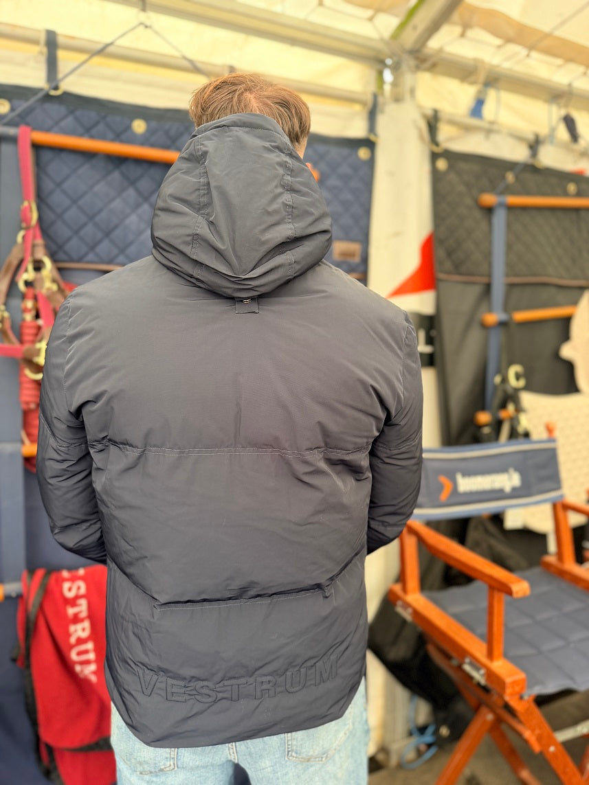 A person viewed from behind wearing a dark gray puffer jacket with a hood, standing inside a tent or workshop with orange chairs and gear in the background.
