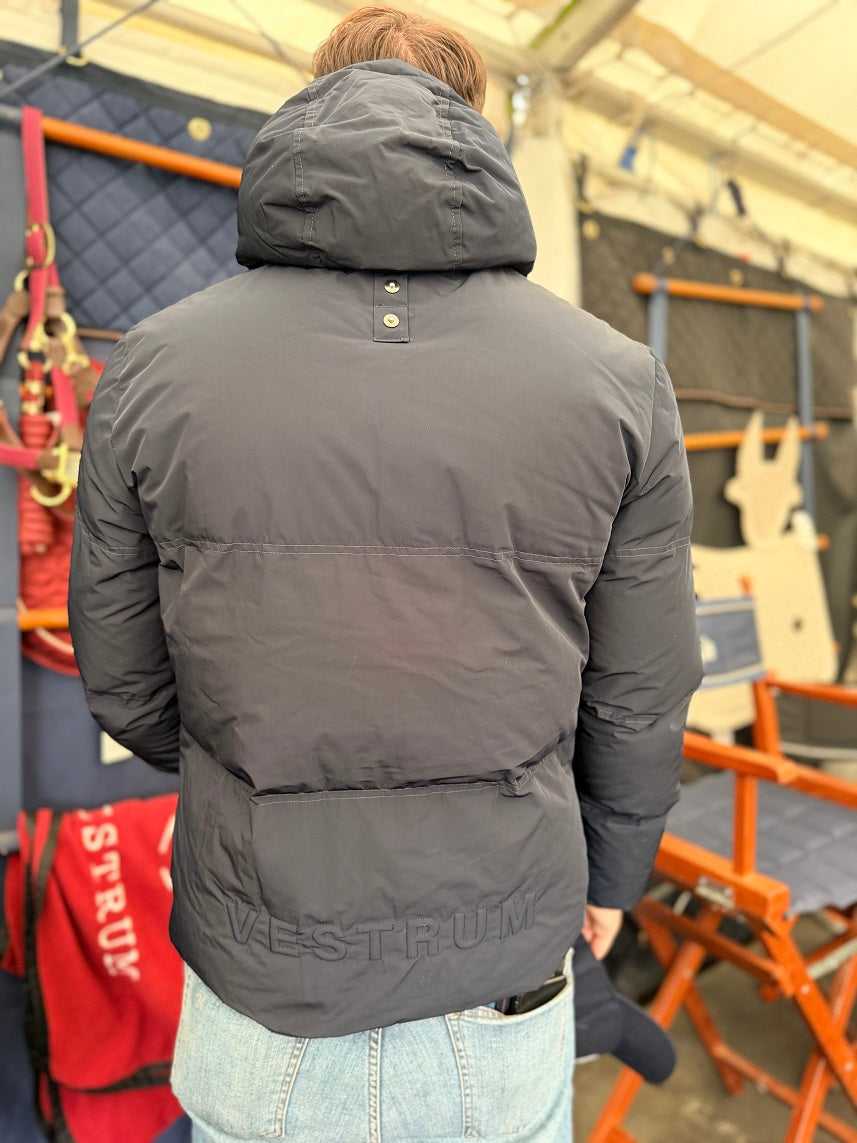 A person viewed from behind wearing a dark gray puffer jacket with 'VESTRUM' printed on the back, paired with light blue jeans in an indoor setting with orange chairs and gear.