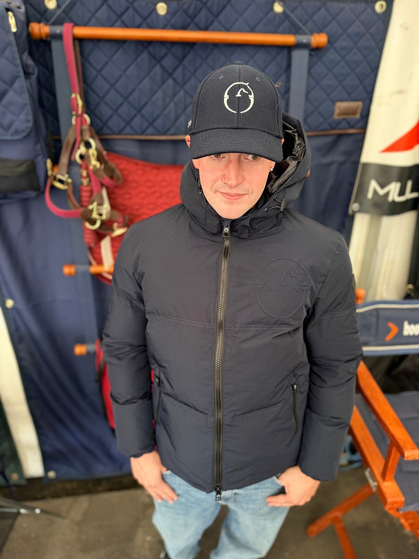 A man wearing a dark navy puffer jacket and matching baseball cap with a circular logo, standing in front of a blue quilted tack wall with horse gear and an orange chair.