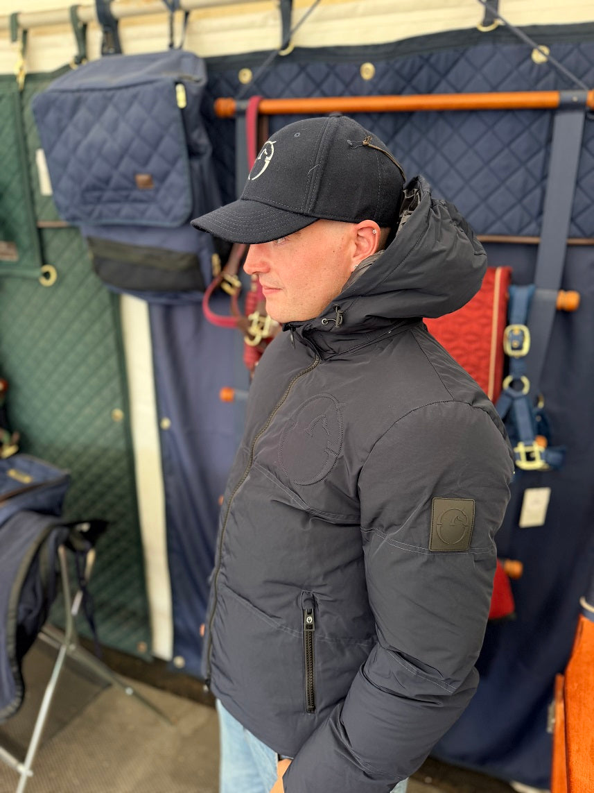 A man wearing a dark gray puffer jacket and a black baseball cap stands in profile inside what appears to be a retail or storage space with hanging bags and gear in the background.