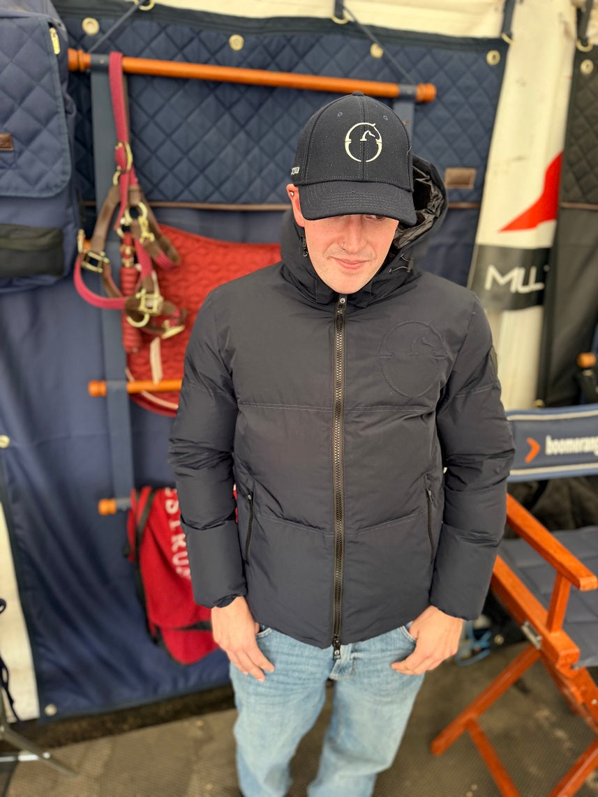 A man wearing a dark gray hooded puffer jacket and a black baseball cap with a white circular logo, standing in front of a blue quilted backdrop with red bags and an orange chair.