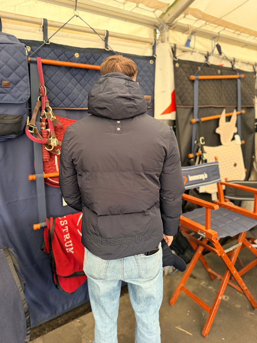 A man viewed from behind wearing a dark gray puffer jacket and light blue jeans, standing inside a tent-like stall surrounded by horse tack, bags, and orange folding chairs.