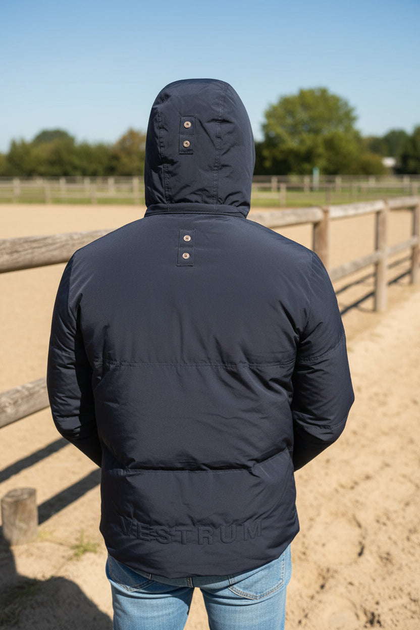 A person viewed from behind wearing a dark gray hooded puffer jacket with a visible Vestrum branding print on the lower back, standing inside what appears to be a tent or storage area with equipment in the background.